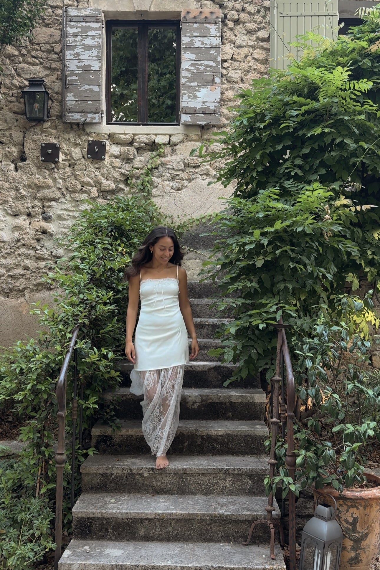 Woman in a light dress standing on stone steps in a garden setting with a stone building in the background.