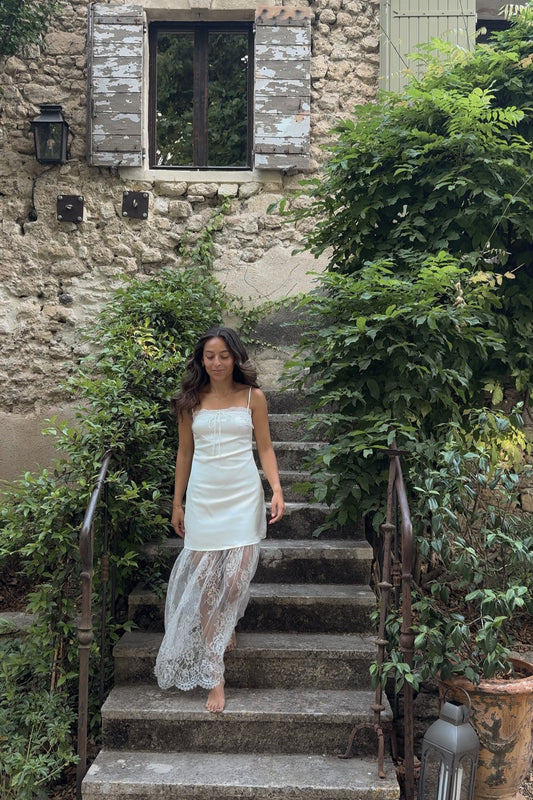 Woman in a light dress standing on stone steps in a garden setting with a stone building in the background.