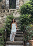 Woman in a light dress standing on stone steps in a garden setting with a stone building in the background.
